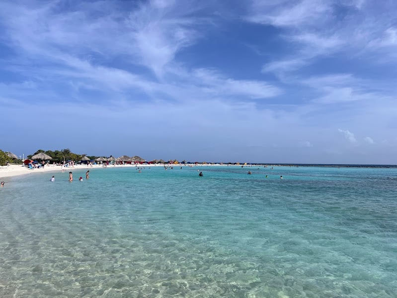 Imagem da praia Baby Beach em Aruba durante o dia com mar a frente com algumas pessoas dentro e ao fundo faixa de areia com guarda-sóis de palha.