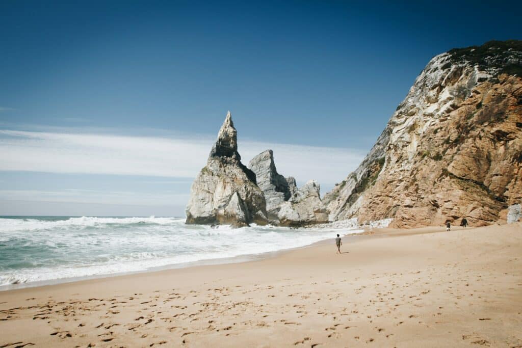 uma pessoa sozinha de costas ao longe em meio à areia fofa que se encontra com um mar calmo e formações rochosas pontudas atrás em Praia da Ursa, em Portugal, que pode ser visitado com o CDAM
