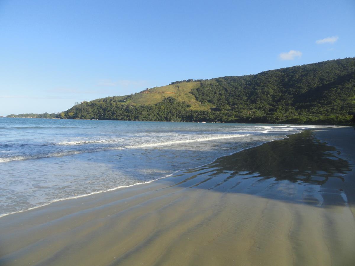 Praia de Maranduba com mar calmo, faixa de areia extensa, no fundo uma montanha verde com árvores e céu azul durante o dia. Imagem ilustrando post Airbnb em Maranduba.