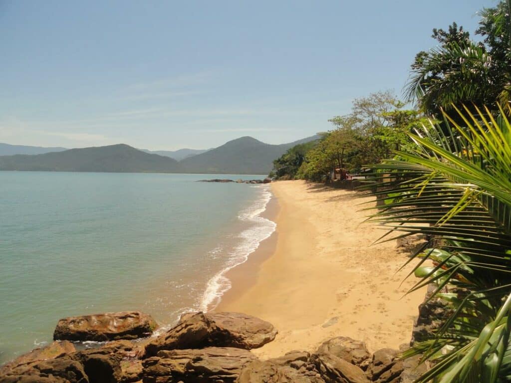 Praia de Picinguaba com uma faixa de areia pequena do lado direito e do lado esquerdo o mar calmo. Em volta tem várias árvores verdes e algumas pedras. Imagem ilustrando post Airbnb em Picinguaba.