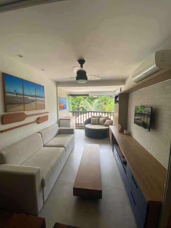 Sala em PRAIA DO FORTE CONDOMÍNIO KAUAI. É bem moderna e conta com um sofá à esquerda, mesa de centro retangular e painel de televisão com várias gavetas. No fundo tem uma varanda com vista e um puff com almofadas.