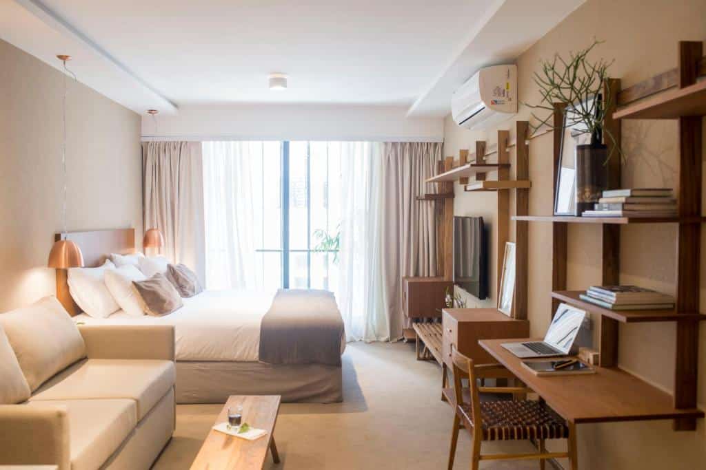 quarto bastante amplo do AQ Tailored Suites com muitas decorações em nichos e prateleiras de parede, mesinha de trabalho com cadeira e tv em frente a uma cama bem grande com mesinha e luminária em ambos os lados, além de um sofá e uma janela ampla do outro lado