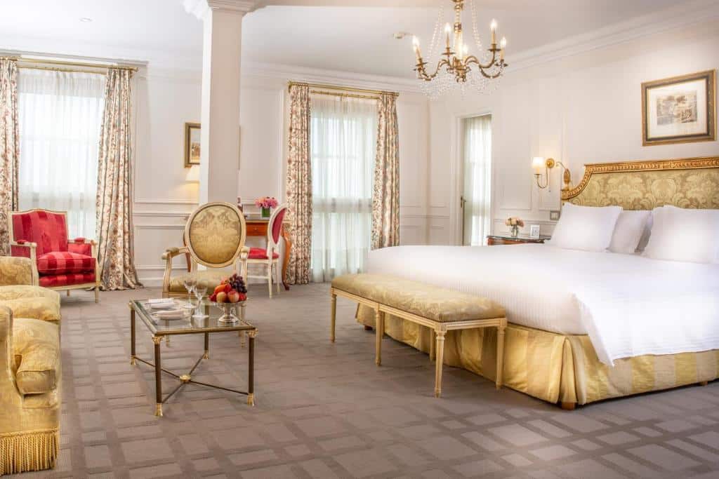 quarto do Alvear Palace Hotel bastante amplo com decoração estilo clássico com detalhes em dourado e vermelho, cama de casal, mesinha de centro de vidro, poltronas, lustre e janelas grandes com cortinas