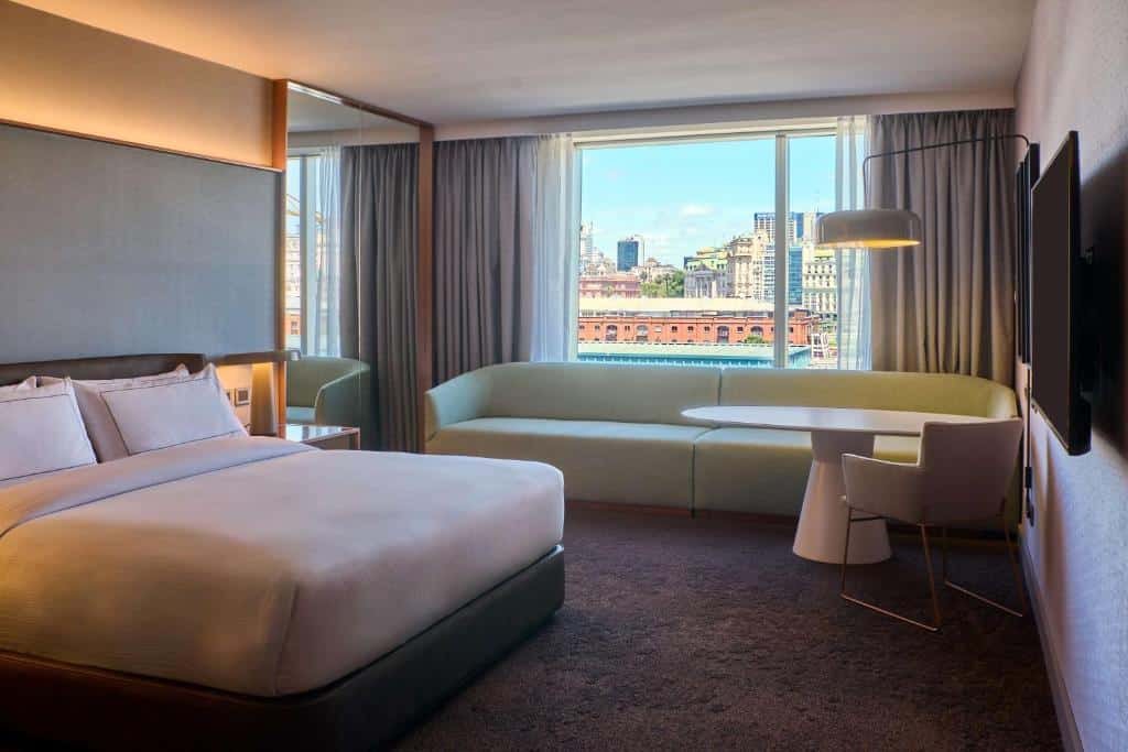 quarto do Hilton Buenos Aires com cama de casal, tv em frentem mesinha e luminária em ambos os lados, além de uma cabeceira com luz atrás, há um sofá comprido com mesinha redonda e poltrona abaixo da janela com vista