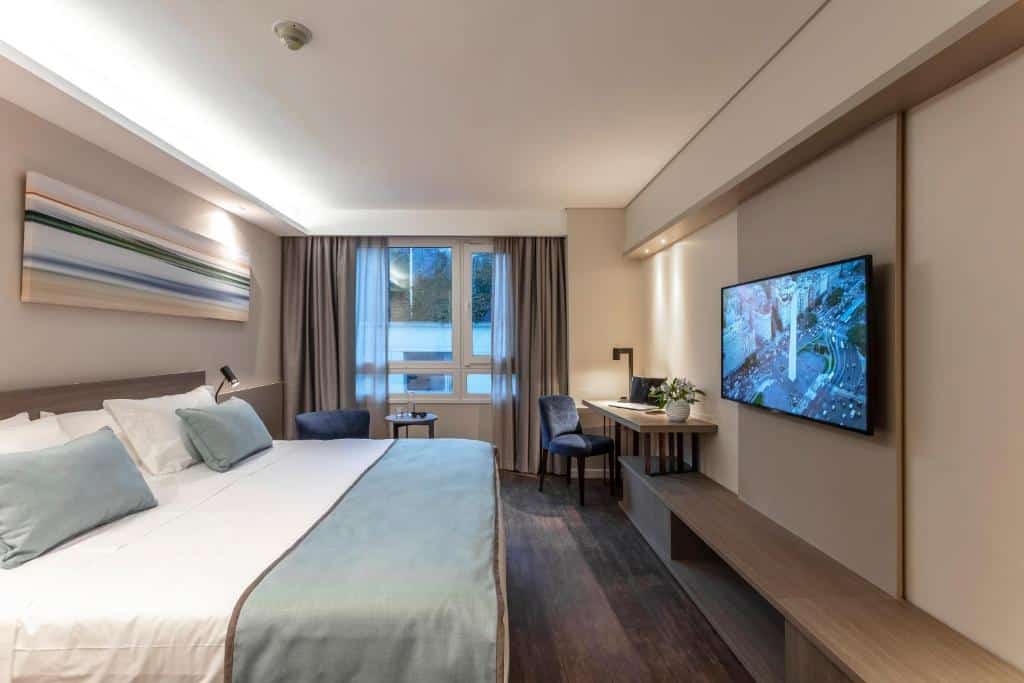 quarto do Huinid Obelisco Hotel com cama de casal bastante grande, tv em frente, mesinha de cabeceira em ambos os lados, luminária nas paredes, há ainda uma mesinha e cadeira e janela com vista e cortinas, o ambiente é bastante amplo