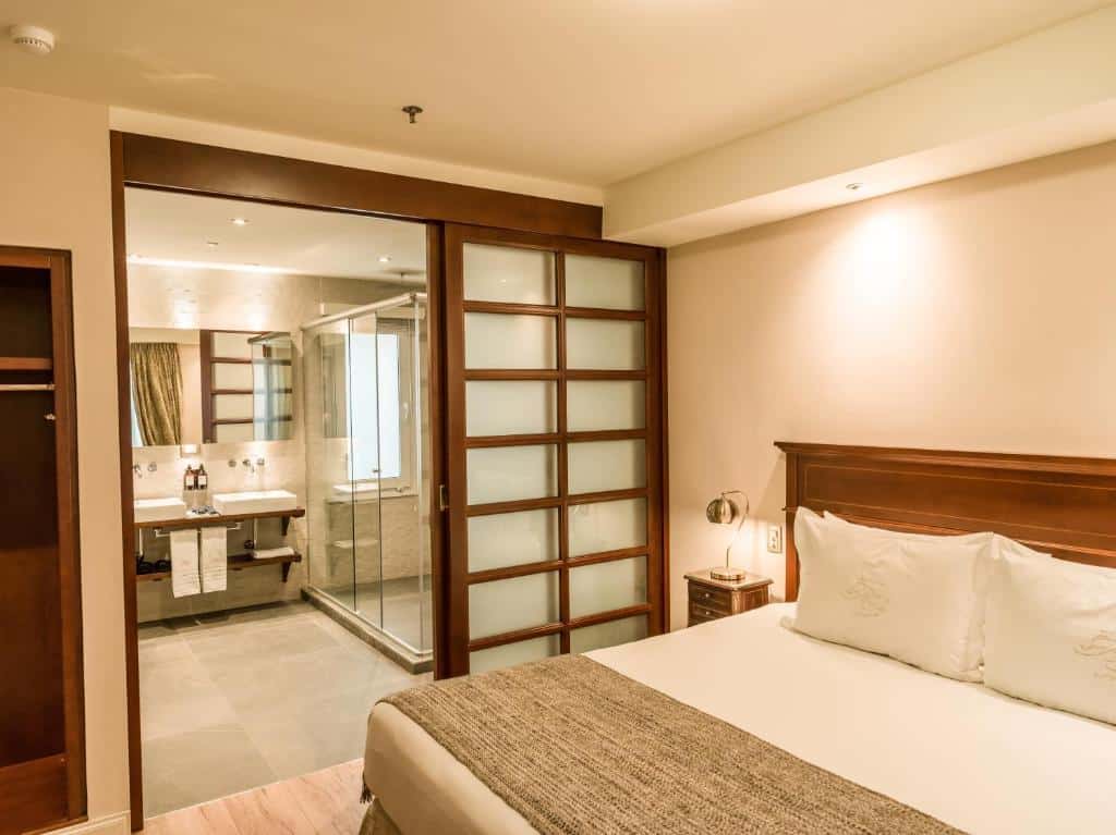 quarto do Melia Recoleta Plaza Hotel com cama de casal, mesinha e luminária, além de um banheiro grande com box e pia ampla, com acesso por uma porta de correr de madeira 