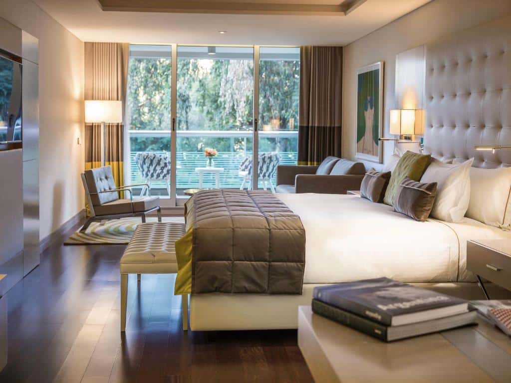 quarto do Palladio Hotel Buenos Aires - MGallery com cama de casal grande, mesinha de ambos os lados com livro em cima, do outro, uma porta grande de vidro para sacada com mesinha e cadeiras do lado de fora, há quadro na parede e luminária na parede, os tons são brancos e marrons