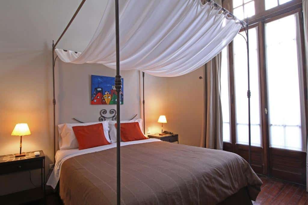 Quarto do A Hotel com uma pequena sacada do lado direito, uma cama de casal, mesinhas de cabeceira com pequenos abajures e um pequeno quadro sob a cama