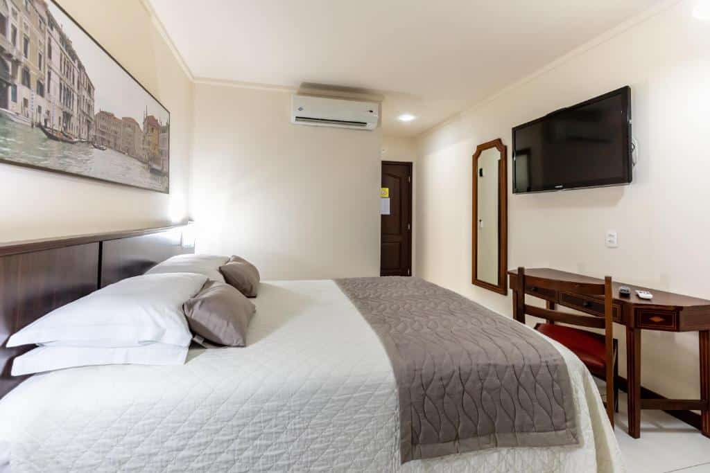 Quarto do Aguas do Iguaçu Hotel Centro com uma cama de casal e em frente dela uma escrivaninha com cadeira, uma tv na parede um um espelho do lado. Do lado esquerdo do quarto tem um ar-condicionado e uma porta ao fundo.