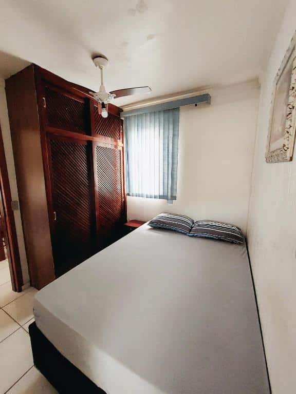 Quarto do Apartamento Ubatuba 150m do mar Maranduba com uma cama de casal encostada na parede direita, uma janela na parede ao fundo e do lado na parede esquerda tem um guarda-roupa de madeira.