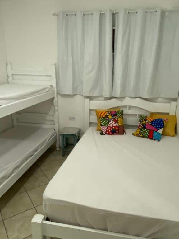 Quarto do Aparthouse Beira Mar com uma cama de casal do lado direito e uma beliche do lado esquerdo. Separando as camas tem um banquinho no chão e uma janela com cortinas na parede.