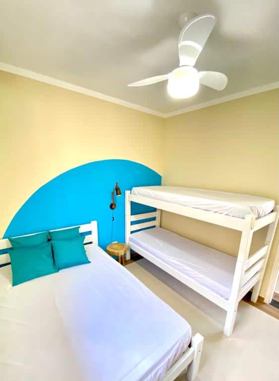 Quarto do Aparthouse Maranduba com uma cama de casal do lado esquerdo e do lado direito uma beliche. Separando as duas camas tem um banquinho e no teto tem um ventilador.