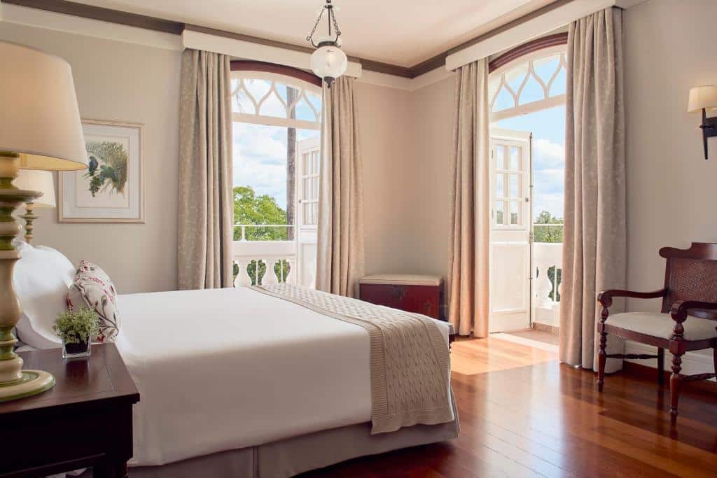 Quarto do Belmond Hotel das Cataratas que mostra uma cama de casal, de cada lado da cama uma mesa de cabeira e em cima um abajur. No quarto há duas portas abertas que dá acesso a sacada com vista para a natureza.