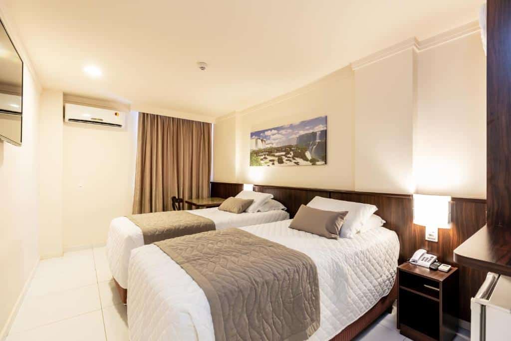 Quarto do Bogari Hotel que mostra duas camas de solteiro lado a lado. Do lado da cama esquerda tem uma mesa de cabeceira e um frigobar. Já do lado da cama direita tem uma mesa, uma cortina e um ar-condicionado.