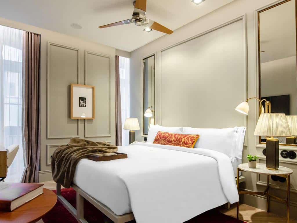 Quarto do Browns Avenue Hotel com uma cama de casal com almofadas e um cobertor, há um ventilador de teto, uma pequena sacada com cortinas, mesinhas de cabeceira com abajures e dois espelhos, para representar hotéis perto da Praça Marquês de Pombal em Lisboa