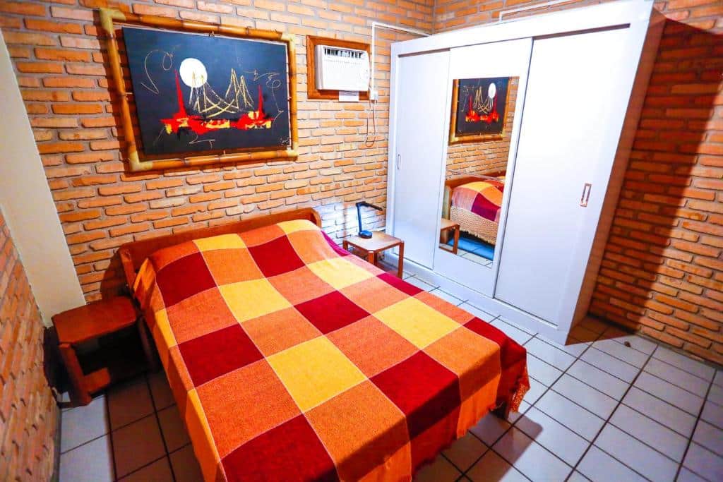Quarto do airbnb Cabanas Jurerê. A direita há um armário, que conta com espelho no meio. No lado esquerdo do quarto há uma cama de casal, com cômodas dos dois lados e abajur em uma delas. Imagem utilizada para ilustrar o post airbnb em Jurerê Internacional.