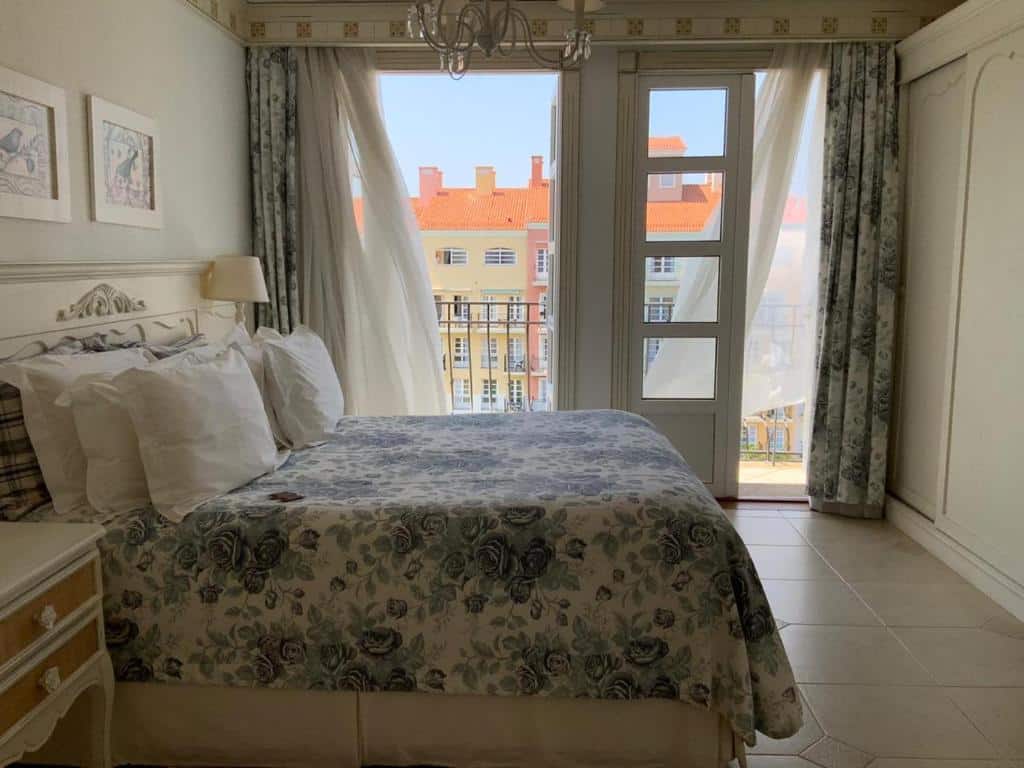 Quarto do airbnb Il Campanario - Cobertura. Cama de casal ao centro do quarto, dos dois lados da cama há cômodas com abajur. Ao fundo há duas portas grandes com cortinas, essas portas levam a varanda do quarto. Imagem utilizada para ilustrar o post airbnb em Jurerê Internacional.