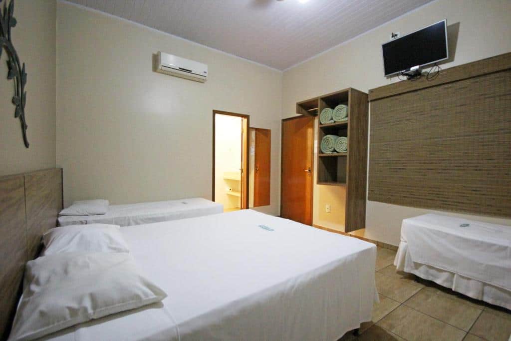 Quarto do Carpe Diem Park Hotel com uma cama de casal, duas de solteiro, uma televisão, um armário com toalhas e uma janela com persiana, para representar hotéis em Olímpia