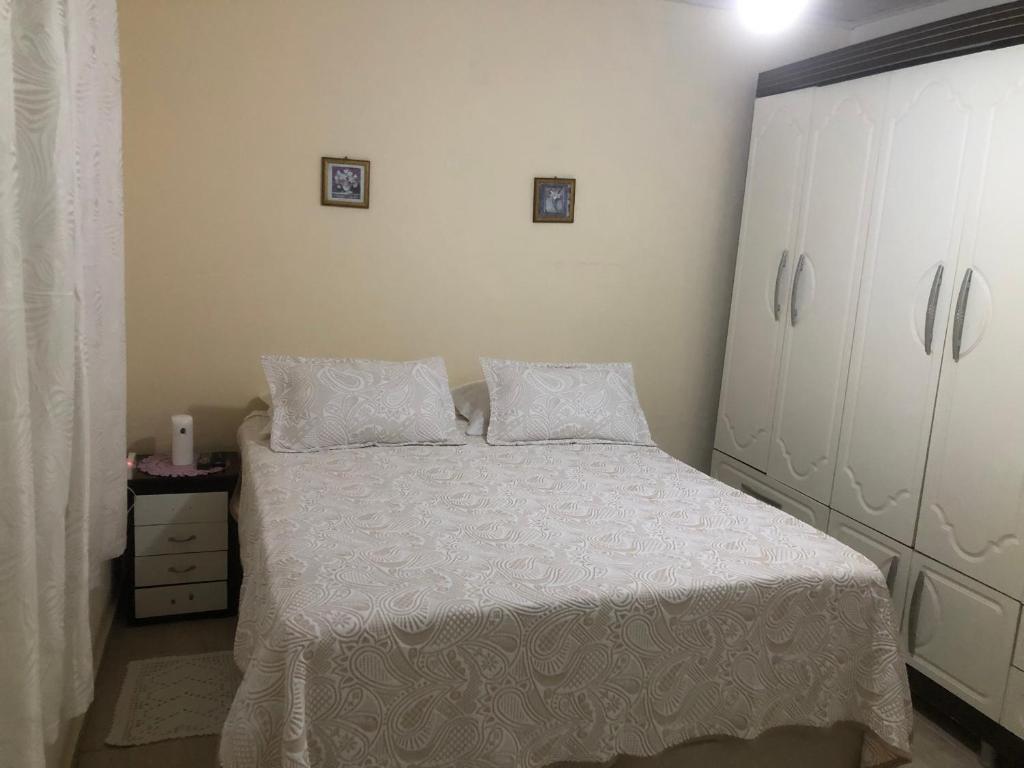 Quarto da Casa Ampla e Confortável com uma cama de casal, do lado direito um guarda-roupa e do lado esquerdo uma mesa de cabeceira e uma janela na parede com cortinas.