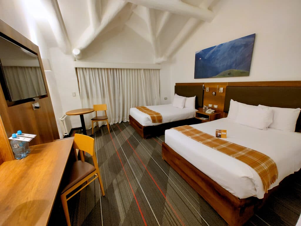 quarto da Casa Andina Premium Valle Sagrado Hotel & Villas com duas camas de casal, dispostas lado a lado, na parte direita da suíte. O chão é revestido de carpete e há duas mesas de madeira na frente das camas