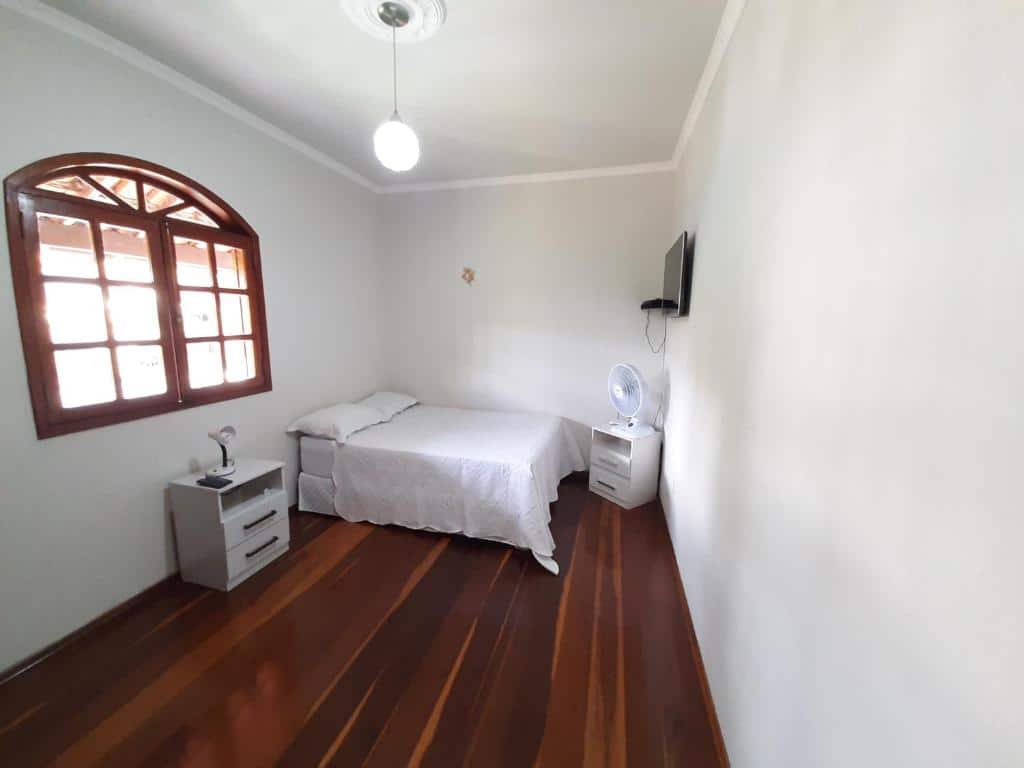 Quarto da Casa da Tuca com cama de casal do lado esquerdo da imagem ao fundo com uma cômoda do lado direito da cama em frente a cama uma cômoda com ventilador e uma TV na parede. Representa airbnb em São João del Rei.
