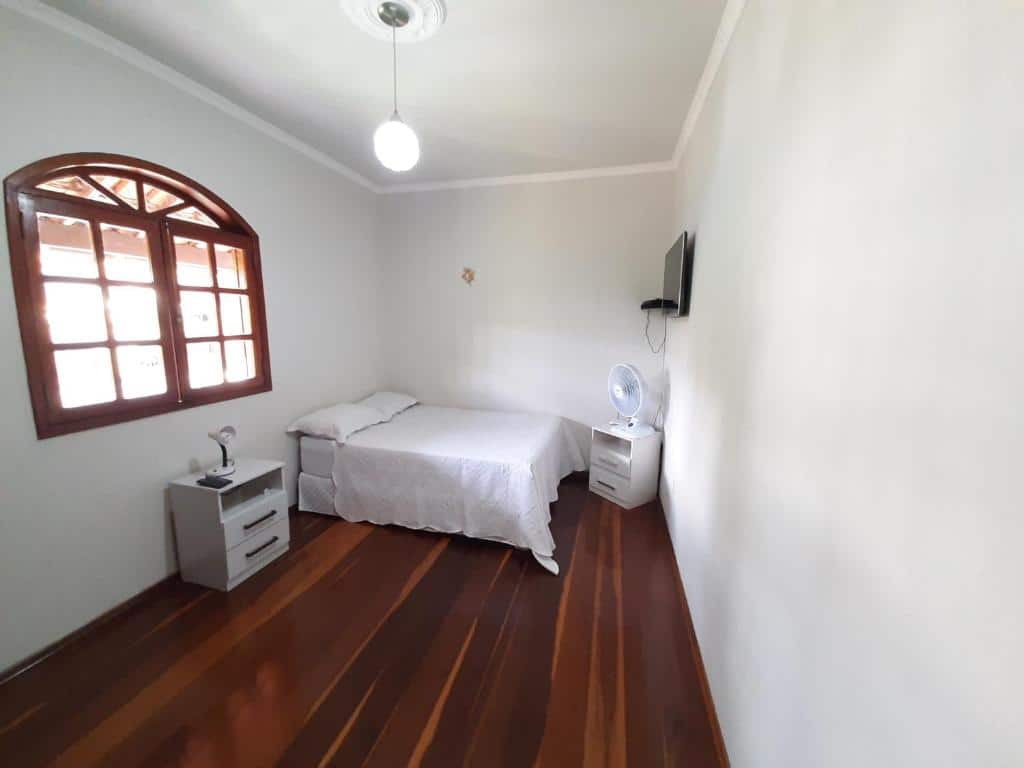 Quarto da Casa da Tuca com cama de casal do lado esquerdo da imagem ao fundo com uma cômoda do lado direito da cama em frente a cama uma cômoda com ventilador e uma TV na parede. Representa airbnb em São João del Rei.