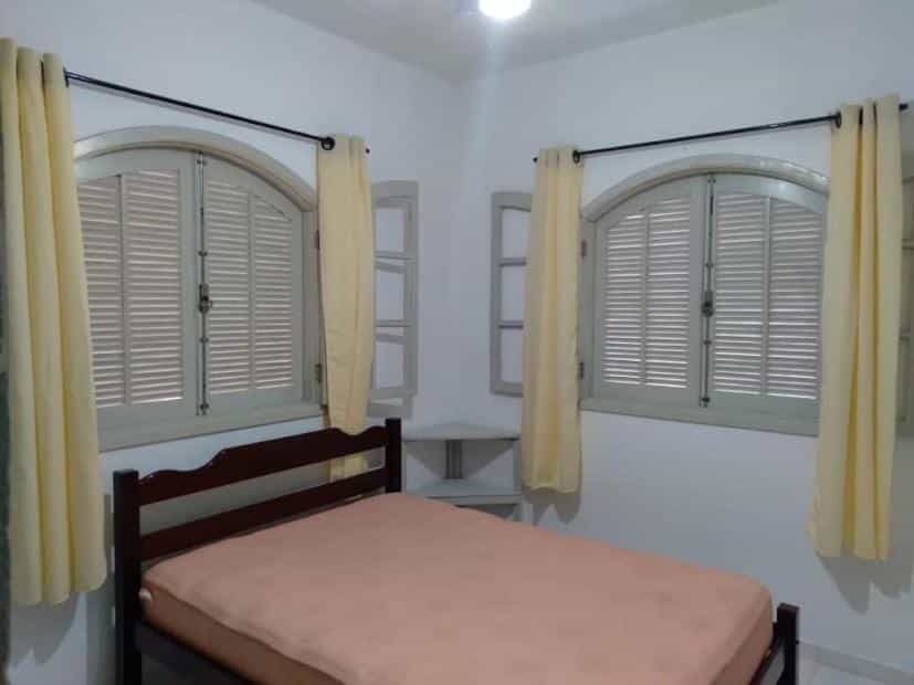 Quarto da Casa Ubatuba, Maranduba, piso superior com uma cama de casal ao centro e uma mesinha de canto. Na parede atrás da cama e na parede do lado esquerdo tem uma janela com cortinas.