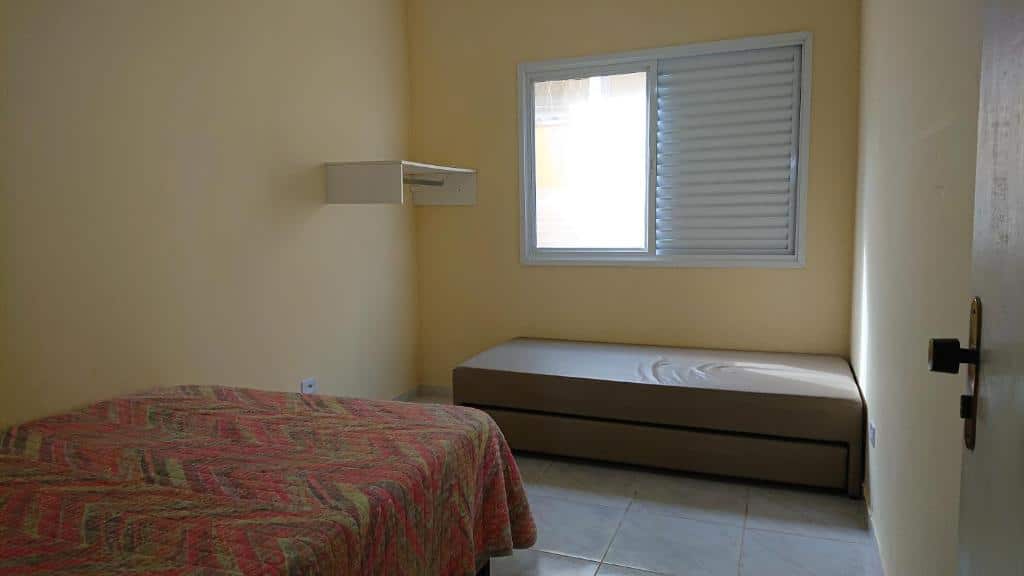 Quarto dos Chalés Mallumar com uma cama de casal no canto esquerdo encostada na parede e ao fundo encostada na parede da janela uma cama de solteiro e em frente uma prateleira na parede.