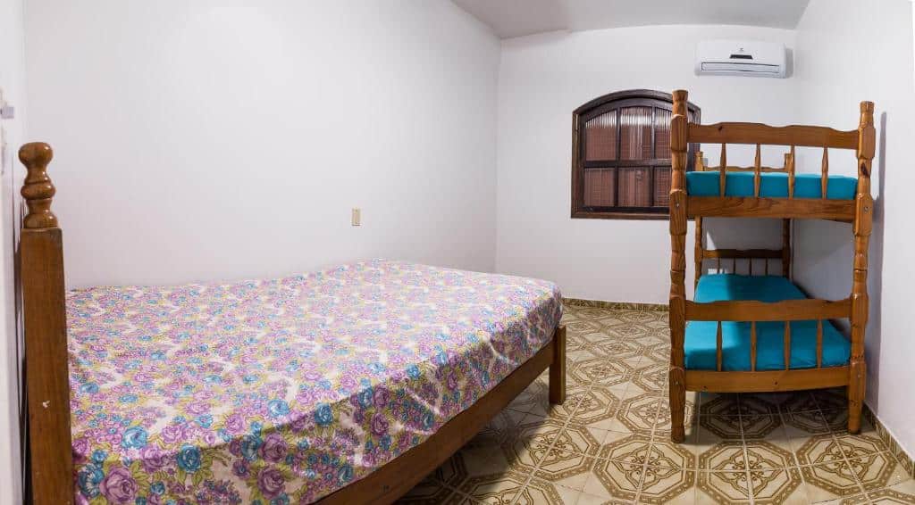 Quarto dos Chalés Pajuçara com uma cama de casal no canto esquerdo e do lado direito do quarto tem um beliche. No quarto também tem uma janela de madeira e um ar-condicionado perto do beliche.