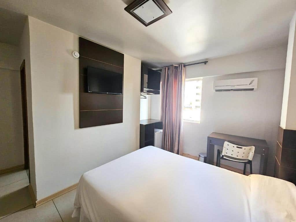Quarto do CLH Suites Foz do Iguaçu que mostra uma cama de casal e de frente uma tv na parede. Do lado direito da cama tem uma escrivaninha com cadeira, um ar-condicionado, uma janela com cortinas, dois armários e um frigobar. 