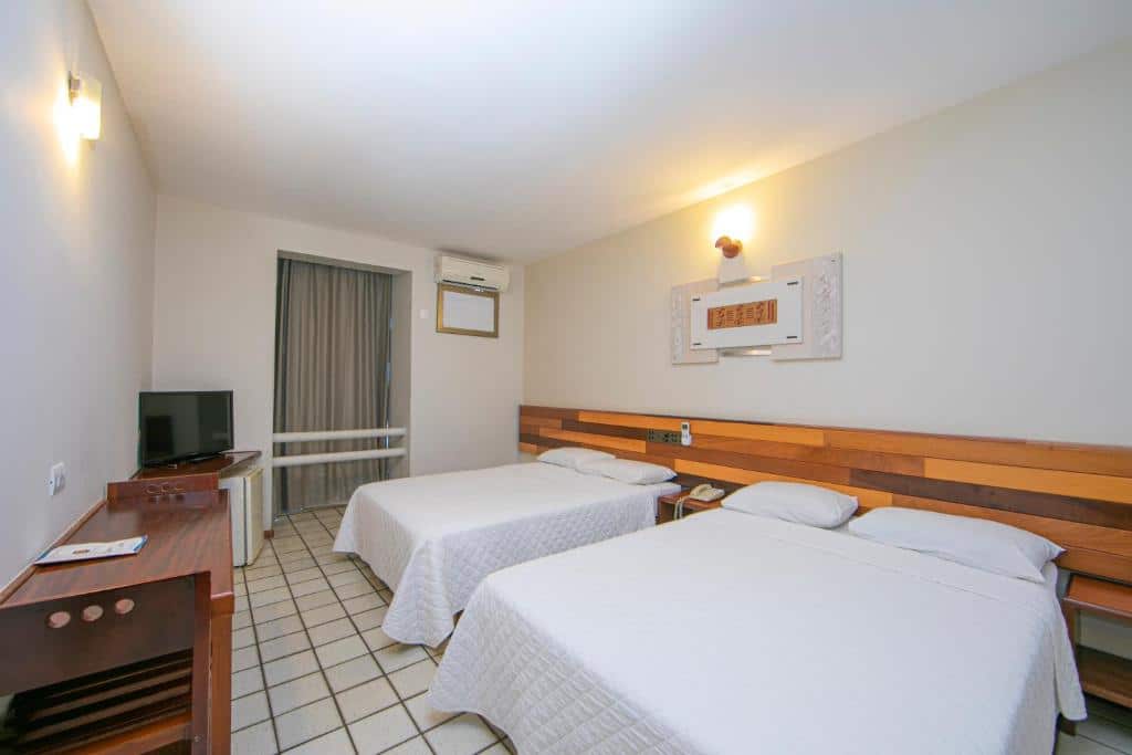 Quarto do Falls Galli Hotel com duas camas de casal, em frente da cama tem uma mesa com duas cadeiras e uma tv em um móvel. Do lado da cama direita tem um ar-condicionado na parede e uma porta com cortinas.