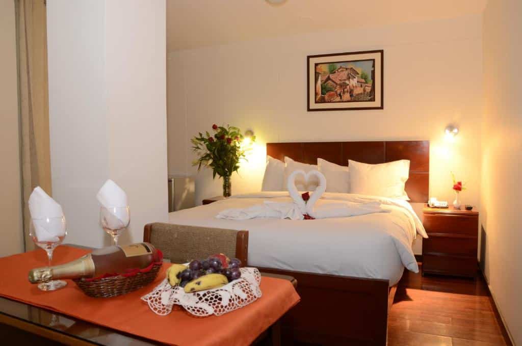 quarto do Golden Inca Hotel com uma cama de casal ao fundo, no centro da imagem, onde duas toalhas estão enroladas e dobradas como dois cisnes em cima da colcha. Em frente a cama há uma mesa com duas taças de vidro e uma garrafa de champanhe.