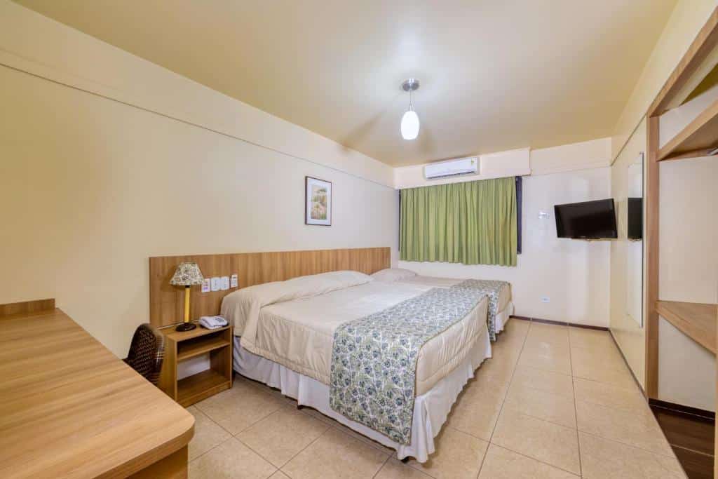 Quarto do Hotel Colonial Iguaçu com uma cama de casal e do lado esquerdo da cama tem uma cama de solteiro. Do lado esquerdo da cama de solteiro tem uma janela com cortinas, um ar-condicionado e uma tv. Já do lado direito da cama de casal tem uma mesa de cabeceira e uma escrivaninha com uma cadeira.