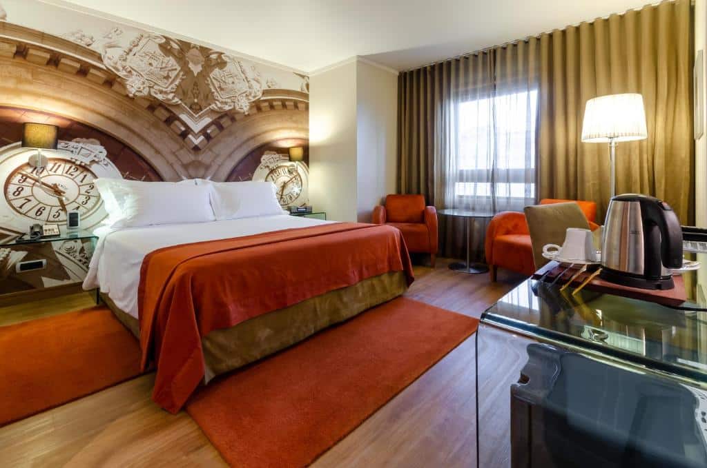 Quarto do Hotel Marques De Pombal com uma cama de casal, do lado direito há uma janela com cortinas e duas poltronas estofadas, em frente a cama, há uma mesa de vidro com comodidades para preparar café, para representar hotéis perto da Praça Marquês de Pombal em Lisboa