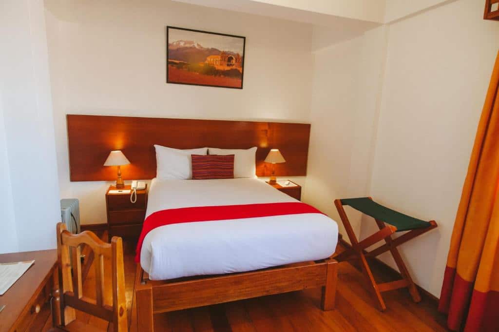 quarto do Hotel Monasterio San Pedro com uma cama de casal pequena, duas cômodas com abajures em cada lado da cama, além de uma banqueta de tecido e uma mesa de madeira em frente a cama.