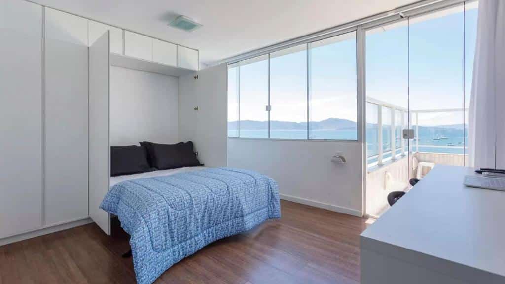 Quarto do airbnb Mergulho no Mar de Jurerê com Vista Frontal. A direita há uma mesa com duas cadeiras, na esquerda é possível observar a cama de casal e armários que a cercam. Ao fundo têm janelas que dão vista para o mar, assim como uma porta que dá acesso a varanda. Imagem utilizada para ilustrar o post airbnb em Jurerê Internacional.