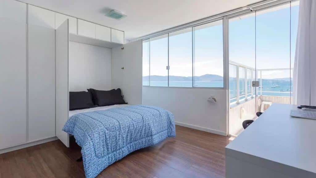 Quarto do airbnb Mergulho no Mar de Jurerê com Vista Frontal. A direita há uma mesa com duas cadeiras, na esquerda é possível observar a cama de casal e armários que a cercam. Ao fundo têm janelas que dão vista para o mar, assim como uma porta que dá acesso a varanda. Imagem utilizada para ilustrar o post airbnb em Jurerê Internacional.
