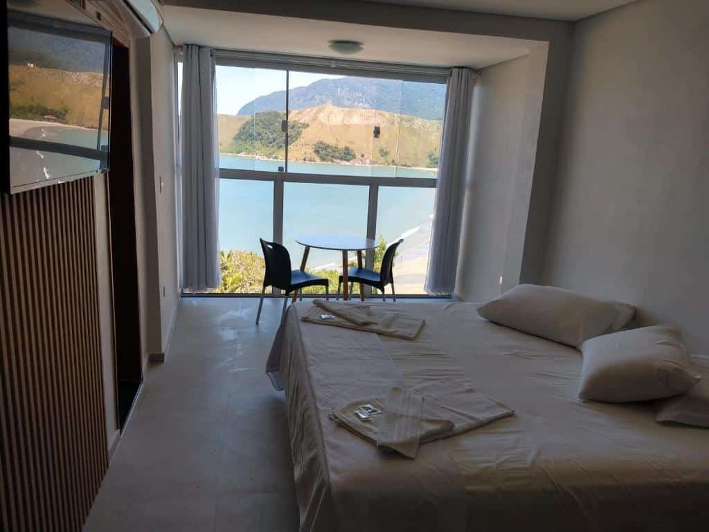 Quarto do Panoramica Residencial com cama de casal do lado direito da imagem, em frente a cama uma parede com TV e do lado direito da cama uma mesa redonda com duas cadeiras com vista para o mar. Representa airbnb na praia de Guaecá.