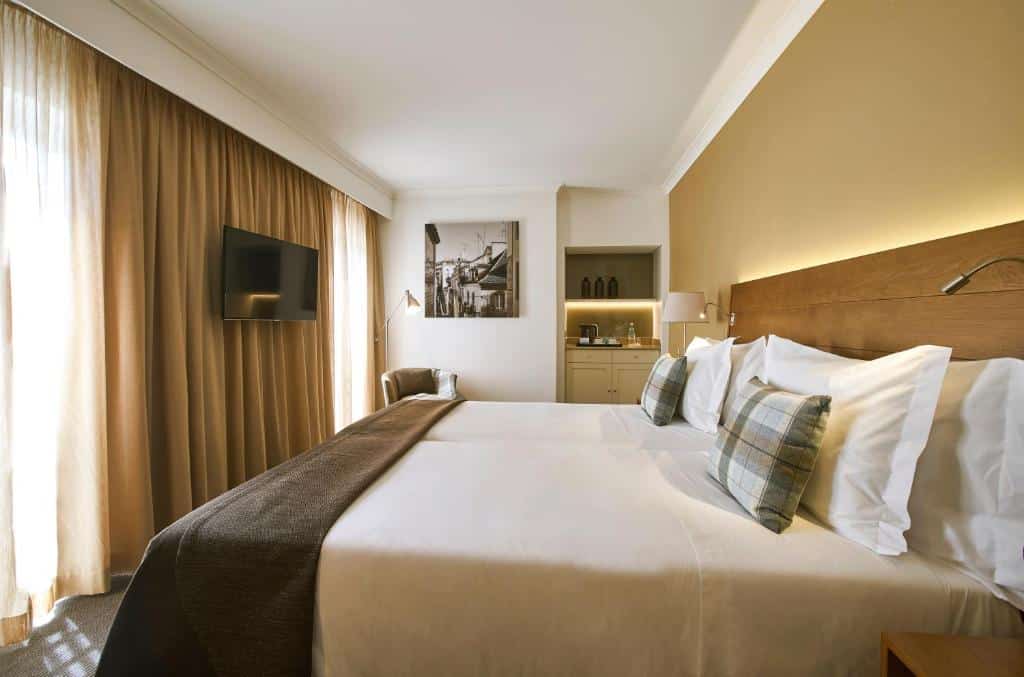 Quarto do PortoBay Marquês com duas janelas com cortinas, próximo delas há uma poltrona e uma luminária de chão, há também uma mini cozinha com alguns itens básicos, no local há uma cama de casal e, de frente para ela, uma televisão.
