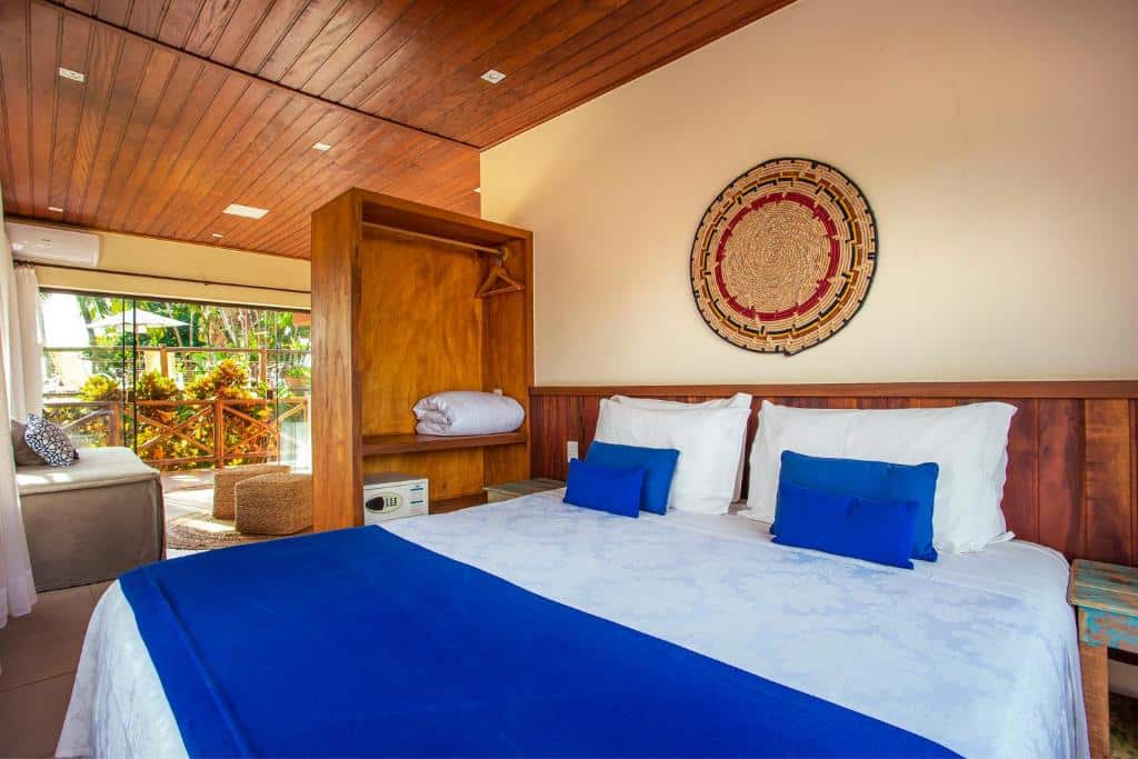 Quarto da Pousada Altamira. Há uma grande cama de casal com um armário e cabideiro. Ao fundo é possível ver uma sala de estar com sofá e alguns banquinhos de palha. Depois da sala há uma grande porta de vidro com cortinas, que vai para a área externa do quarto. Imagem utilizada para ilustrar o post pousadas pet friendly em Ilhabela.