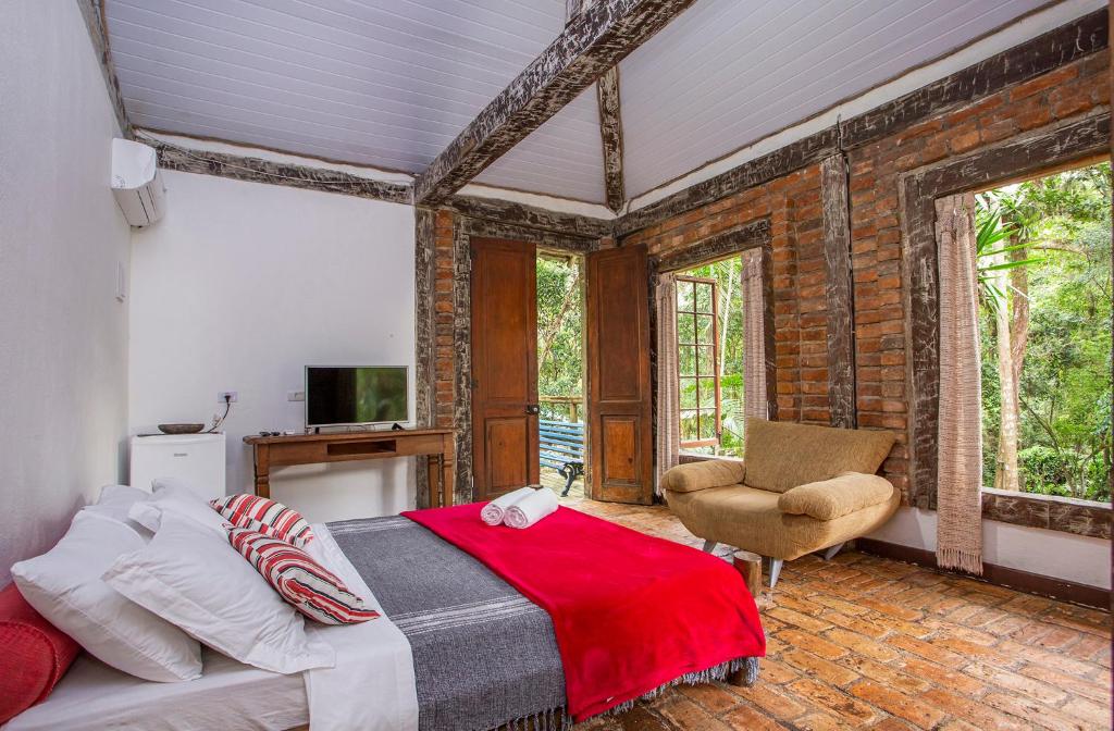 Quarto da Pousada Barulho D' água. Na direita há duas grandes janelas com cortinas e no meio uma grande poltrona. A esquerda há uma cama de casal. No fundo do quarto há uma televisão e um frigobar, além de uma grande porta dupla de madeira que vai para a varanda do quarto. Imagem utilizada para ilustrar o post pousadas pet friendly em Ilhabela.