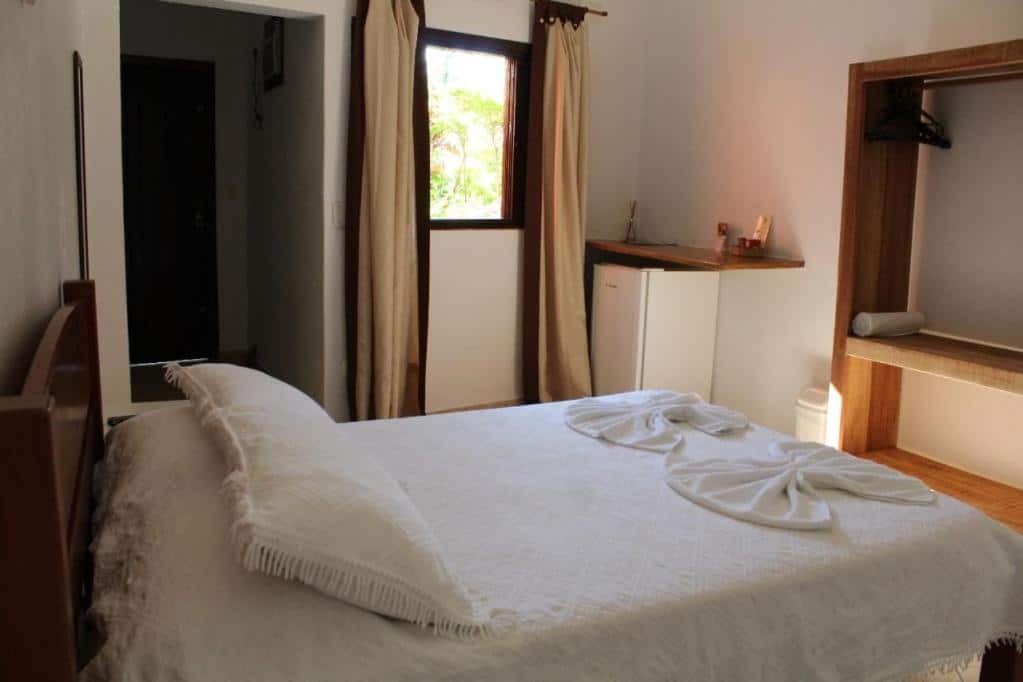 Quarto da Pousada Cantinho D'Abrantes. Na direita há um cabideiro feito em madeira, além de um frigobar com prateleira acima. Na esquerda há uma cama de casal. Ao fundo está uma pequena janela com cortinas. Imagem utilizada para ilustrar o post pousadas pet friendly em Ilhabela.