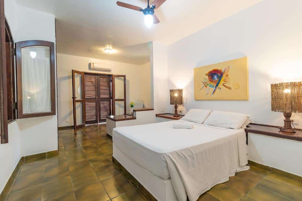 Quarto da Pousada Casa Amarela. Na direita há uma cama de casal com prateleiras e abajures dos dois lados, a esquerda está uma janela. Ao fundo do quarto é possível ver um frigobar e uma porta dupla de madeira que está fechada. Imagem utilizada para ilustrar o post pousadas pet friendly em Ilhabela.