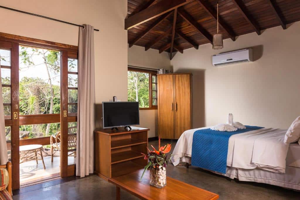 Quarto da Pousada Ilhote da Prainha. A direita é possível ver uma cama de casal, um armário de duas portas e uma janela com vista para a floresta. Na esquerda observa-se uma televisão em cima de rack de madeira, a frente da televisão uma pequena mesa baixa com planta em cima e uma porta com cortinas, que vai para a varanda. Imagem utilizada para ilustrar o post pousadas pet friendly em Ilhabela.