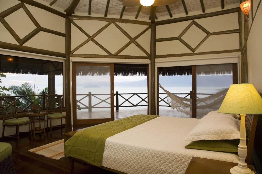 Quarto da Pousada Singuitta. Na direita há uma cama de casal com abajur ao lado, na esquerda algumas cadeiras e uma pequena mesa. Ao fundo há portas que vão para a área externa do quarto, que têm rede, parapeito e vista para o mar. Imagem utilizada para ilustrar o post pousadas pet friendly em Ilhabela.