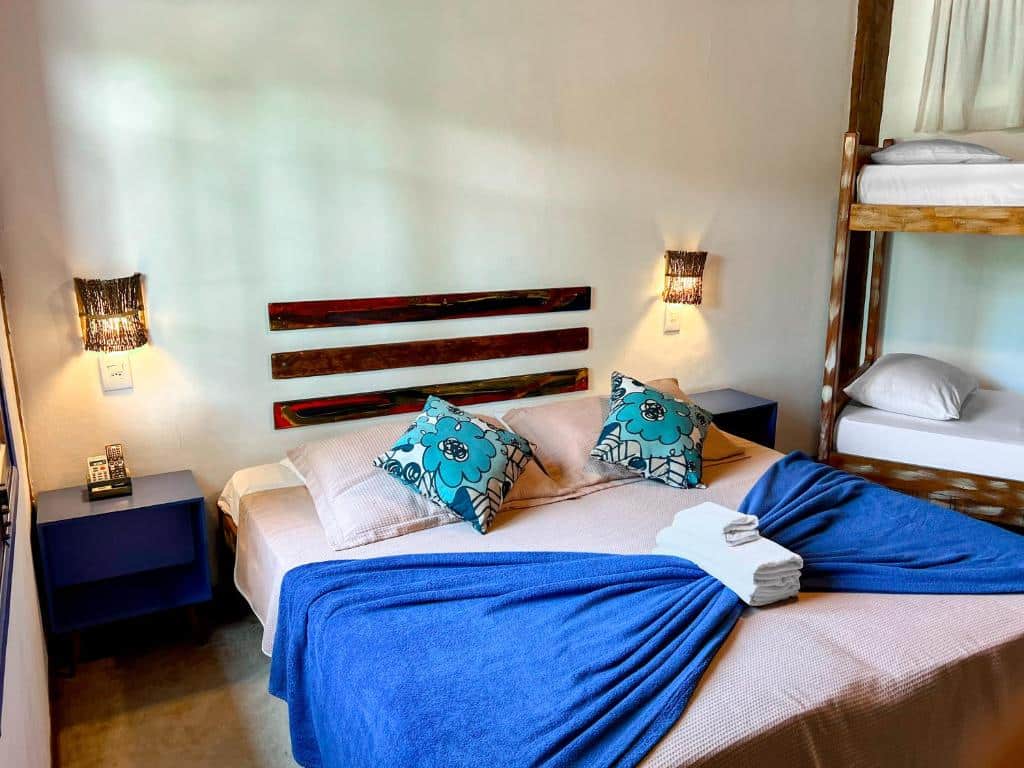 Quarto da pousada Vila Pequeá. Ao centro há uma cama de casal com cômodas e abajures dos dois lados. No lado direito é possível ver um beliche. Imagem utilizada para ilustrar o post pousadas pet friendly em Ilhabela.