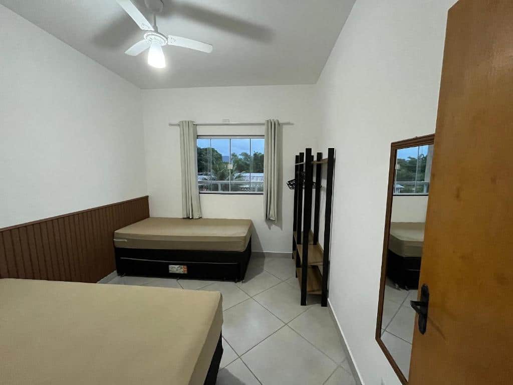 Quarto do Rafael Apartamentos 800m da Praia da Maranduba com duas cama de casal com cabeceiras de madeira, um espelho, uma arara para colocar roupas e objetos e ao fundo uma janela com cortinas.