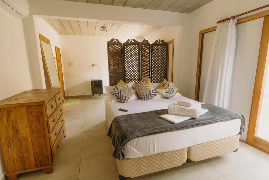 Quarto da pousada Reserva Ilhabela. Na direita há uma cortina, ao centro uma cama de casal e a esquerda uma cômoda de madeira com gavetas, ao fundo é possível ver um biombo de madeira separando o ambiente em dois. Imagem utilizada para ilustrar o post pousadas pet friendly em Ilhabela.