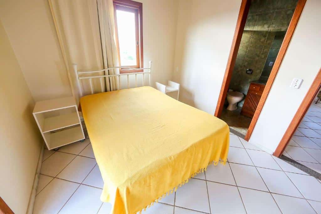 Quarto do airbnb Residencial Casa Tomate é Destino Floripa. Na direita há uma porta que vai para o banheiro da suíte, no centro do quarto está uma cama de casal, com cômodas dos dois lados. Acima da cama há uma janela com cortina. Imagem utilizada para ilustrar o post airbnb em Jurerê Internacional.