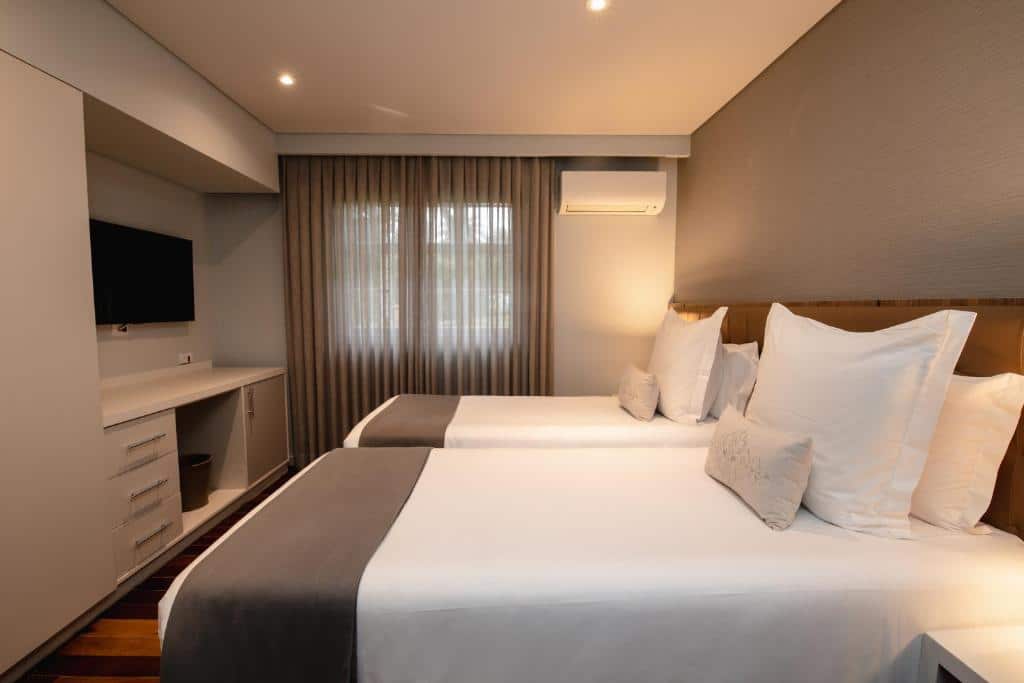 Quarto do Sanma Hotel com duas camas de solteiro uma do lado da outra e cabeceira de madeira. Do lado da cama direita tem um ar-condicionado e uma janela com cortina. Já na frente das camas tem uma tv com um pequeno armário embaixo.